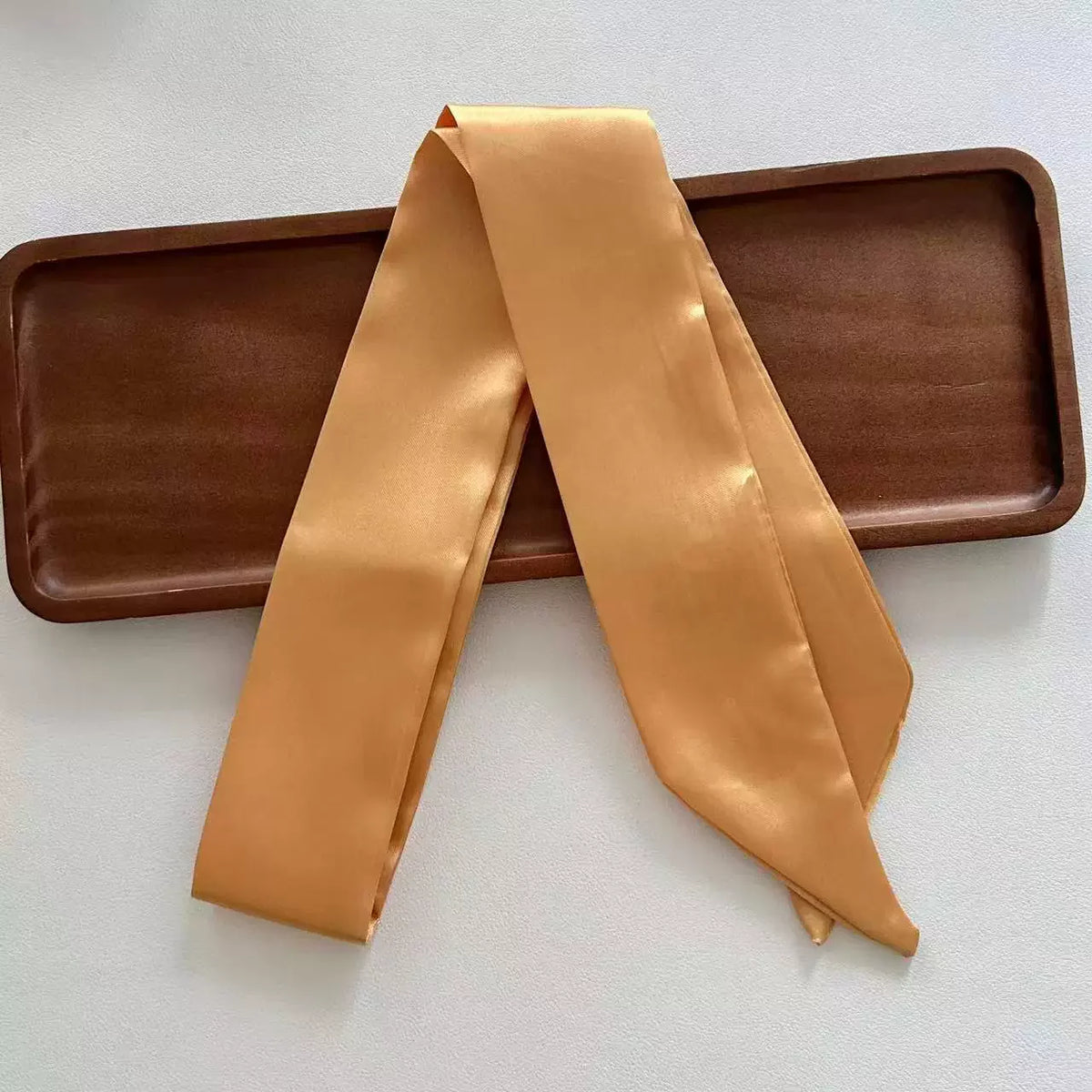 Khaki skinny satin ribbon scarf draped over a wooden tray on a white background
