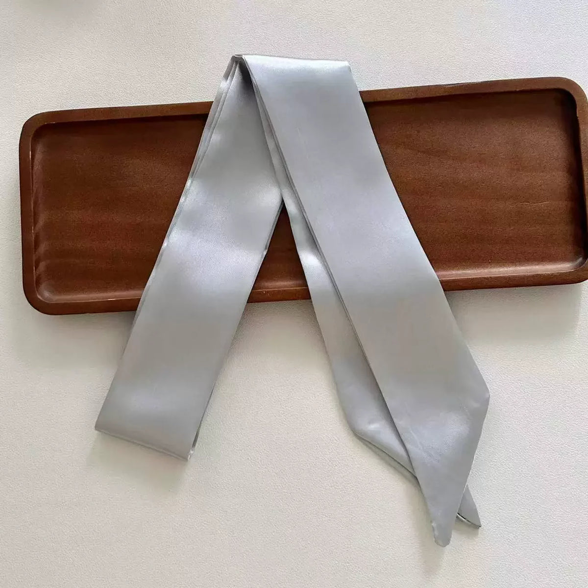 Light gray skinny satin ribbon scarf on a wooden tray with a plain background